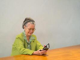 ein Alten asiatisch Frau tragen Brille mit ein Smartphone und lächelnd während Sitzung auf ein Stuhl mit Raum zum Text. Konzept von alt Menschen und Kommunikation foto