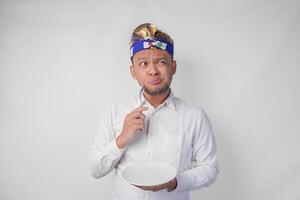 nachdenklich jung balinesisch Mann im Weiß Hemd und traditionell Kopfschmuck halten ein leeren Teller mit Kopieren Raum und Löffel während Denken Was Essen zu Essen foto