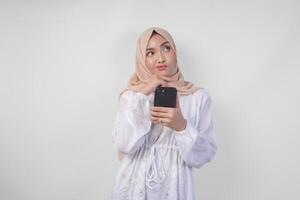 nachdenklich jung asiatisch Muslim Frau tragen Weiß Kleid und Kopftuch, mit Smartphone während halten ihr Kinn und Denken mit ernst Ausdruck Über isoliert Weiß Hintergrund foto