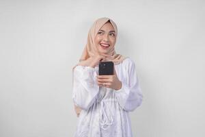 nachdenklich jung asiatisch Muslim Frau tragen Weiß Kleid und Hijab halten Telefon während Denken und finden Idee mit glücklich Ausdruck, isoliert auf Weiß Hintergrund. Ramadan und eid Mubarak Konzept foto