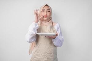 glücklich jung asiatisch Muslim Frau im Hijab und Sahne Schürze halten ein leeren Teller mit Kopieren Raum während Herstellung ein köstlich Hand Geste zu ausdrücken Wie gut das Essen ist foto