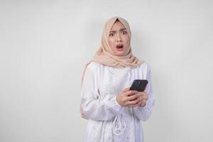 jung asiatisch Muslim Frau tragen Weiß Kleid und Hijab zeigen schockiert Ausdruck auf ihr Gesicht während halten Smartphone, überrascht nach lesen Nachrichten oder Tratsch, isoliert durch Weiß Hintergrund foto