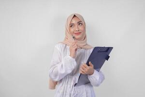 nachdenklich jung asiatisch Muslim Frau tragen Weiß Kleid und Hijab halten ein dokumentieren, Denken während suchen zu das Kopieren Raum über, isoliert durch Weiß Hintergrund. Ramadan und eid Mubarak Konzept foto