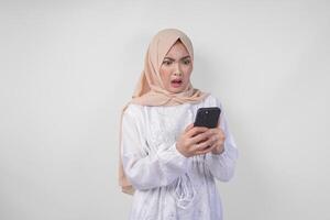 jung asiatisch Muslim Frau tragen Weiß Kleid und Hijab zeigen schockiert Ausdruck auf ihr Gesicht während halten Smartphone, überrascht nach lesen Nachrichten oder Tratsch, isoliert durch Weiß Hintergrund foto