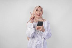 nachdenklich jung asiatisch Muslim Frau tragen Weiß Kleid und Hijab halten Telefon während Denken und finden Idee mit glücklich Ausdruck, isoliert auf Weiß Hintergrund. Ramadan und eid Mubarak Konzept foto