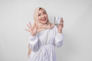 jung asiatisch Muslim Frau im Weiß Kleid und Hijab halten und Trinken Tasse von frisch Wasser während Herstellung ein Okay, Gut, nett Zeichen Geste mit Finger foto