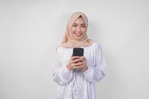 aufgeregt asiatisch Muslim Frau tragen Weiß Kleid und Hijab halten ihr Smartphone während lächelnd fröhlich, isoliert durch Weiß Hintergrund. Ramadhan und eid Mubarak Konzept foto