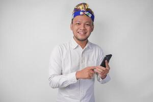 aufgeregt balinesisch Mann tragen Weiß Hemd und traditionell Kopfschmuck lächelnd glücklich während mit Smartphone, antworten Nachricht, lesen Nachrichten foto