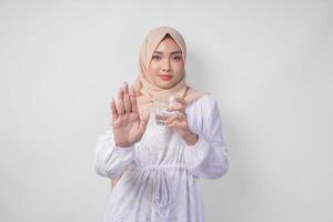ernst jung asiatisch Muslim Frau tragen Weiß Kleid und Hijab zeigen ihr Palme zu machen ein halt Geste während halten ein Tasse von Wasser, Stehen Über isoliert Weiß Hintergrund. Ramadan Konzept foto