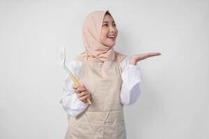 glücklich jung asiatisch Muslim Frau im ein Schleier Hijab und Sahne Schürze halten Spatel während zeigen zu das Kopieren Raum neben ihr foto