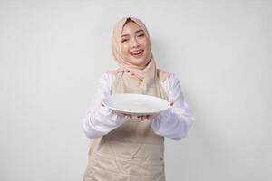 schön jung asiatisch Muslim Frau im Hijab und Sahne Schürze präsentieren ein leeren Teller mit Kopieren Raum Über Es, zeigen das Essen Speisekarte zum iftar foto