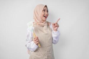 glücklich jung asiatisch Muslim Frau im ein Schleier Hijab und Sahne Schürze halten Spatel während zeigen zu das Kopieren Raum neben ihr foto