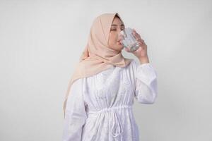durstig jung asiatisch Muslim Frau im Weiß Kleid und Sahne Schleier Hijab Trinken frisch Mineral Wasser nach Fasten. Ramadan Konzept foto