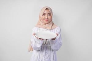 schön jung asiatisch Muslim Frau im Hijab präsentieren ein leeren Teller mit Kopieren Raum Über Es, zeigen das Essen Speisekarte zum iftar foto