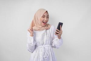jung asiatisch Muslim Frau mit ein glücklich erfolgreich Ausdruck tragen Weiß Kleid und Hijab halten Smartphone, isoliert durch Weiß Hintergrund. Ramadan und eid Mubarak Konzept foto