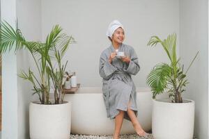 glücklich attraktiv asiatisch Frau im grau Bademantel posieren beim das Badewanne, lächelnd fröhlich während Trinken Tee. Urlaub Freizeit Konzept. foto