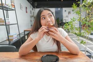 schön asiatisch jung Frau Essen köstlich Gebäck genannt Cromboloni beim ein Café. das Dame beißt Stück von Schokolade Füllung Cromboloni, suchen glücklich beim das Cafe foto
