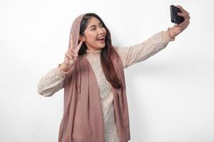 schön asiatisch Muslim Frau im Kopftuch Hijab Schleier Lachen und nehmen ein Spaß Selfie mit Handy, Mobiltelefon Telefon, isoliert durch Weiß Hintergrund foto