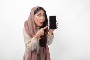 aufgeregt jung asiatisch Muslim Frau im Kopftuch Schleier Hijab zeigen zu das Kopieren Raum beim Smartphone leer Bildschirm, isoliert auf Weiß Hintergrund Studio. Ramadan und eid Mubarak Konzept. foto