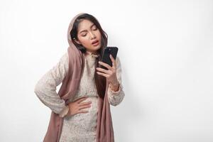 jung asiatisch Muslim Frau im Schleier Hijab Gefühl hungrig während halten ihr Bauch und mit ein Smartphone zu bestellen Lebensmittel. Ramadan und eid Mubarak Konzept foto