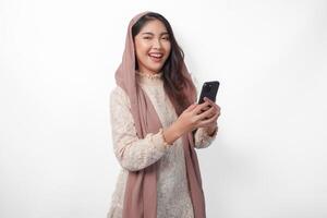 Porträt von jung asiatisch Muslim Frau im Schleier Hijab lächelnd fröhlich während halten und mit ein Smartphone. Ramadan und eid Mubarak Konzept foto