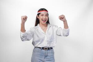 Porträt von ein aufgeregt jung asiatisch Frau erziehen Faust oben Geste, gewinnen und feiern Sieg Pose, tragen Flagge Stirnband und Weiß Shirt. Unabhängigkeit Tag Konzept foto