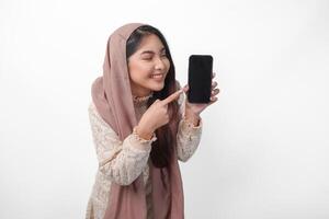aufgeregt jung asiatisch Muslim Frau im Kopftuch Schleier Hijab zeigen zu das Kopieren Raum beim Smartphone leer Bildschirm, isoliert auf Weiß Hintergrund Studio. Ramadan und eid Mubarak Konzept. foto