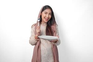 aufgeregt asiatisch Muslim Frau halten Teller und Besteck zu Essen nach Fasten während lächelnd fröhlich auf isoliert Weiß Hintergrund. Ramadan Konzept foto