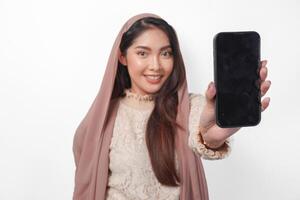 lächelnd jung asiatisch Muslim Frau im Kopftuch Schleier Hijab präsentieren oder zeigen Kopieren Raum auf Smartphone leer Bildschirm, isoliert durch Weiß Hintergrund Studio. Ramadan und eid Mubarak Konzept. foto