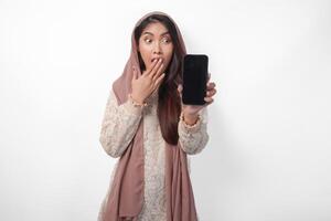 schockiert asiatisch Muslim Frau im Kopftuch Schleier Hijab mit Hand Abdeckung Mund während zeigen leer Telefon Bildschirm mit ein Kopieren Raum, isoliert Weiß Hintergrund foto