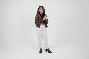 Porträt von ein schockiert asiatisch Frau tragen beiläufig Outfit halten und zeigen ihr Telefon während ihr Mund breit offen, Stehen auf isoliert Weiß Hintergrund foto