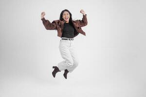 aktiv asiatisch Mädchen ist Springen und erziehen Hände oben tragen braun Jacke, isoliert Weiß Farbe Hintergrund foto
