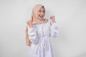 überglücklich jung asiatisch Muslim Frau tragen Hijab und Weiß Kleid Zusammenpressen Faust Gewinner Geste, isoliert auf Weiß Hintergrund. Ramadan und eid Mubarak Konzept foto