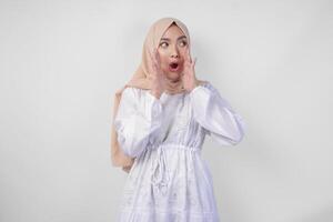 Porträt von jung asiatisch Muslim Frau tragen Weiß Kleid und Hijab mit überrascht Ausdruck und Mund breit öffnen nach Hören schockierend Nachrichten Über isoliert Weiß Hintergrund foto