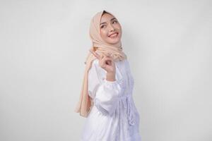 lächelnd asiatisch Frau tragen Hijab und Weiß Kleid zeigen oben zu das Kopieren Raum auf ihr richtig Seite, Stehen Über isoliert Weiß Hintergrund. Ramadan und eid Mubarak Werbung Konzept foto