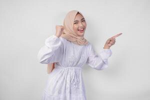überglücklich jung asiatisch Muslim Frau tragen Hijab und Weiß Kleid Zusammenpressen Faust Gewinner Geste während zeigen zu das Kopieren Raum neben ihr, isoliert auf Weiß Hintergrund foto
