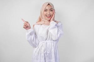 aufgeregt asiatisch Frau tragen Hijab und Weiß Kleid zeigen zu das Kopieren Raum auf ihr richtig Seite, Stehen Über isoliert Weiß Hintergrund. Ramadan und eid Mubarak Werbung Konzept foto