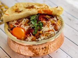 indisch Hähnchen Biryani mit Tomate, Koriander, Cashew Nuss und Brot dum serviert im Kupfer Topf Gericht isoliert auf hölzern Tabelle oben Aussicht Arabisch würzig Essen foto