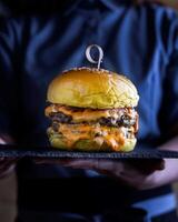 Besondere doppelt Decker Burger mit geschmolzen Käse und Grüner Salat Blätter serviert im Schneiden Tafel im halten Hand isoliert auf hölzern Tabelle Seite Aussicht von Arabisch Essen foto