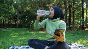 glücklich Muslim Frau Hör mal zu Musik- auf Handy, Mobiltelefon Telefon beim Park während Trinken Mineral Wasser, kostenlos Raum foto