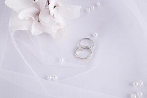 schick Hochzeit Hintergrund im Weiß Töne. zwei Platin Engagement Ringe, ein Diamant Ring auf ein klassisch Weiß Braut- Schleier mit frisch Blumen. oben Sicht. foto