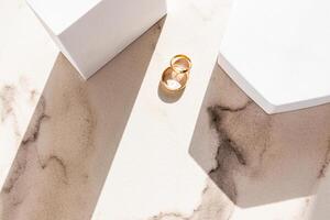 zwei klassisch Gold Hochzeit Ringe Lüge auf ein Marmor Beige Hintergrund zwischen zwei Weiß geometrisch Podeste von zahlen und Sonne Strahlen. oben Sicht. foto