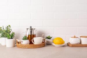 schön Küche Hintergrund mit Glas Teekanne von duftend Tee, Tasse, Zitrone auf Korbweide Tablett. Vorderseite Sicht. Weiß Backstein Mauer. ein Kopieren Raum. foto