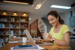 asiatisch Frau Sitzung und halten Anerkennung Karte zum online Einkaufen durch Laptop und Clever Telefon beim heim. foto
