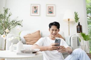 glücklich asiatisch Mann Sitzung auf Couch mit Smartphone im Leben Zimmer beim heim, entspannen Zeit und Lebensstil Konzept foto