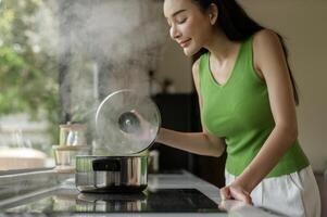 asiatisch Frau Kochen und riechen Verkostung Suppe im ein Topf im das Küche Tabelle beim heim. foto