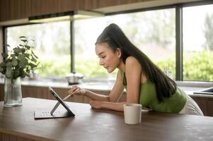 jung asiatisch Frau Arbeiten mit Tablette im das Küche beim Zuhause foto