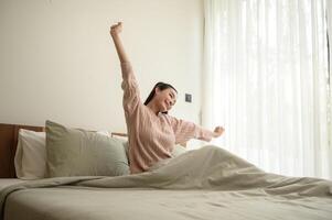 jung asiatisch Frau Dehnen im Bett nach aufwachen oben im das Morgen foto