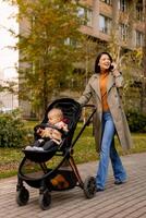 jung Frau mit süß Baby Mädchen im Baby Kinderwagen mit Handy, Mobiltelefon Telefon beim das Herbst Park foto