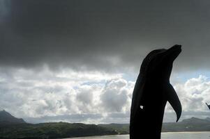 Silhouette von ein Delfin Statue gegen ein Hintergrund von Strand Hügel mit wolkig Wetter foto